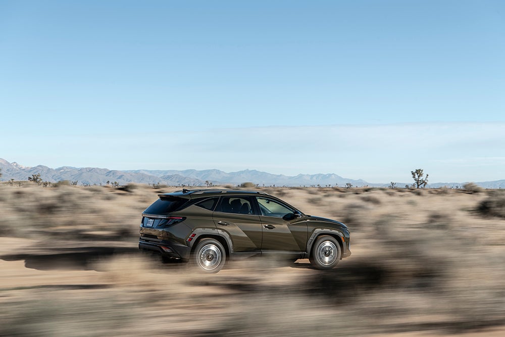 A brown Hyundai Tucson drives quickly across a dusty, arid landscape with mountains in the background.