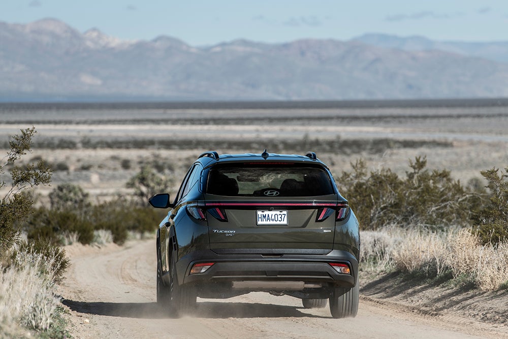 A rear view of a dark green Hyundai Tucson driving down a dirt road, kicking up a small cloud of dust. The vehicle has a prominent horizontal taillight bar, and a mountain range is visible in the distance under a bright sky.