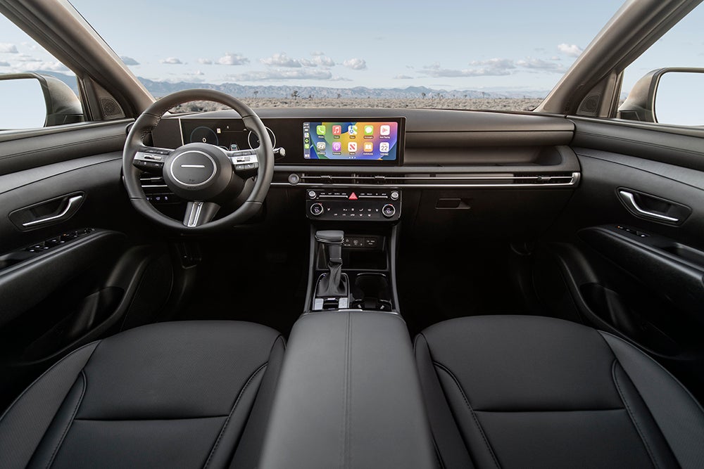 A front-on view of the interior of a Hyundai, showcasing the dashboard, steering wheel, infotainment screen, and black leather seats. The view out the windshield shows a distant mountain range under a cloudy sky.
