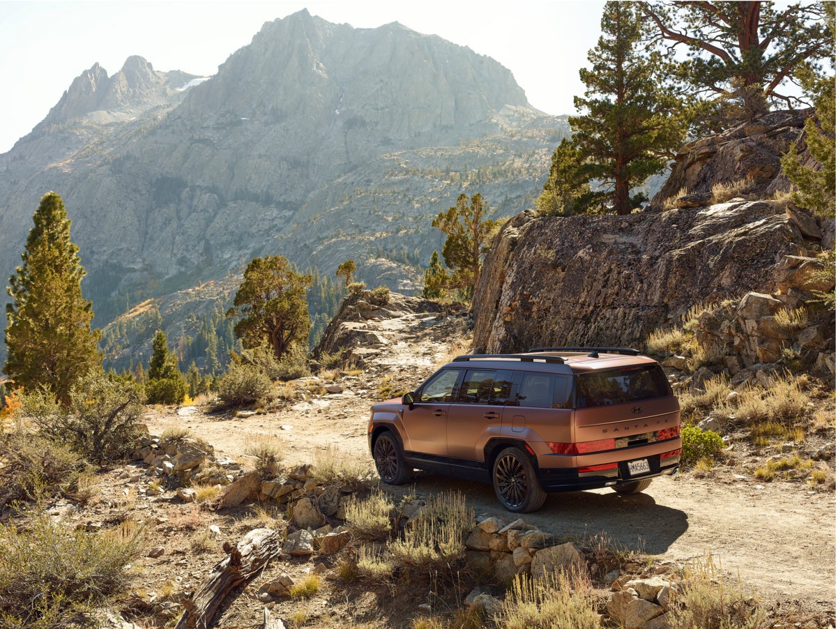 A brown 2026 Santa Fe Hybrid Santa Fe Hybrid in Phoenix drives away from the viewer to the left seen from a high angle on a rocky trail next to a stone mountain face overlooking a tree-filled valley.