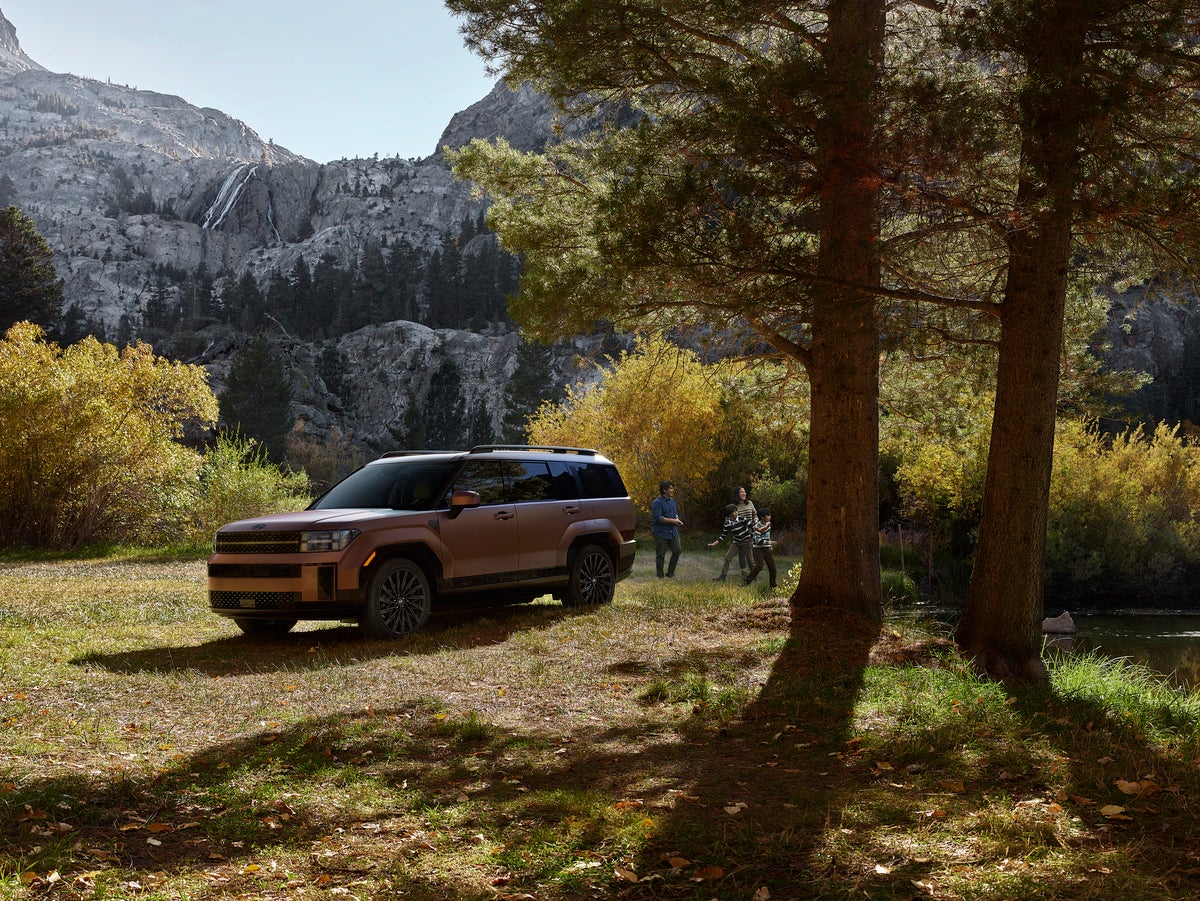 A brown new Santa Fe Hybrid in Phoenix is parked facing the viewer to the left on a grass field next to a small river with a family laughing and playing to the right of the vehicle and a tree-covered mountain in the background.