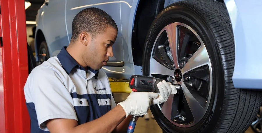 A male auto mechanic in a red, white, and navy blue uniform uses a pneumatic impact wrench to tighten or loosen the lug nuts on a car tire. The vehicle is raised on a red hydraulic lift inside a brightly lit professional garage.